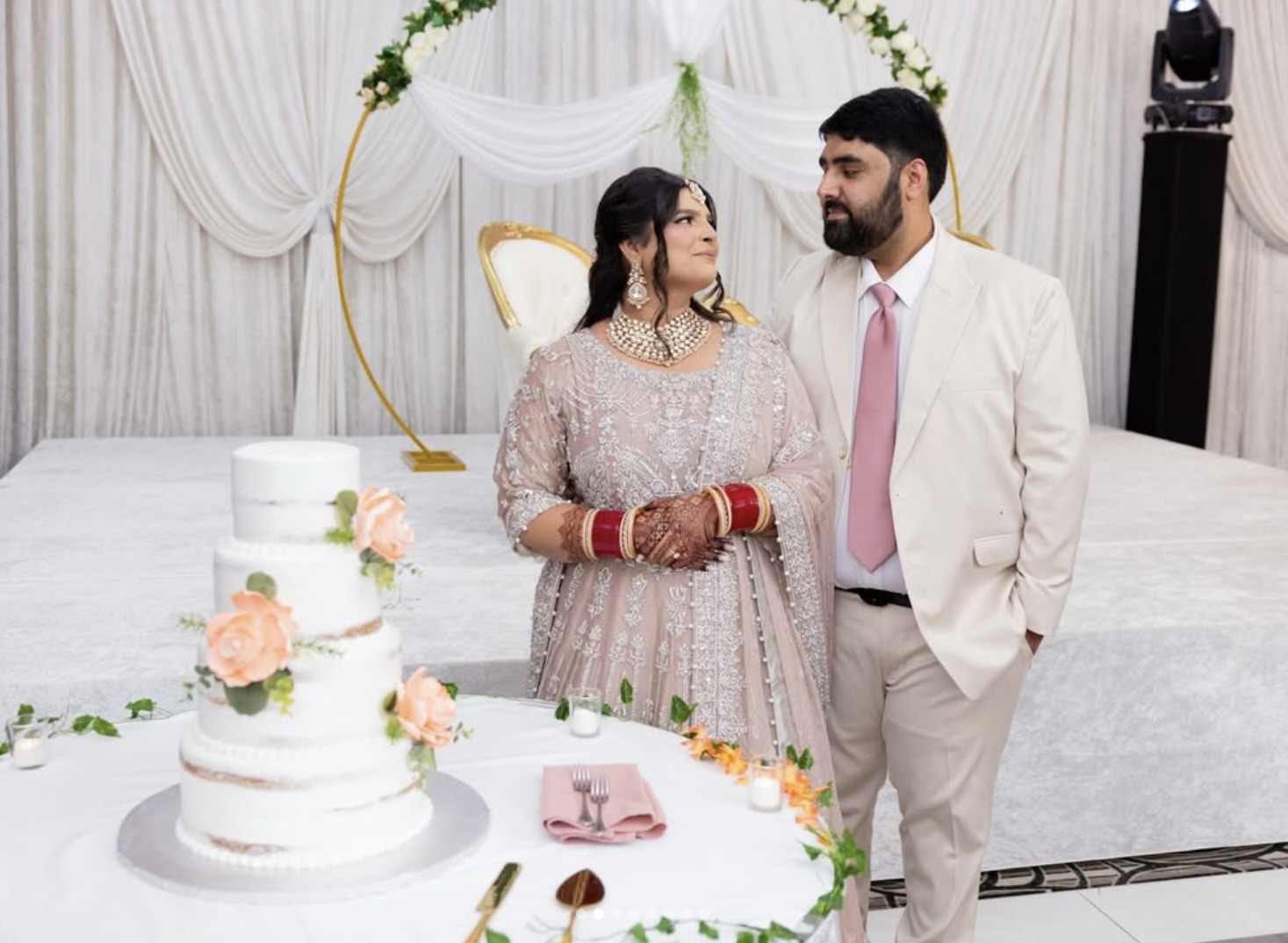 Bride and groom beside their tiered wedding cake under a floral arch at the reception